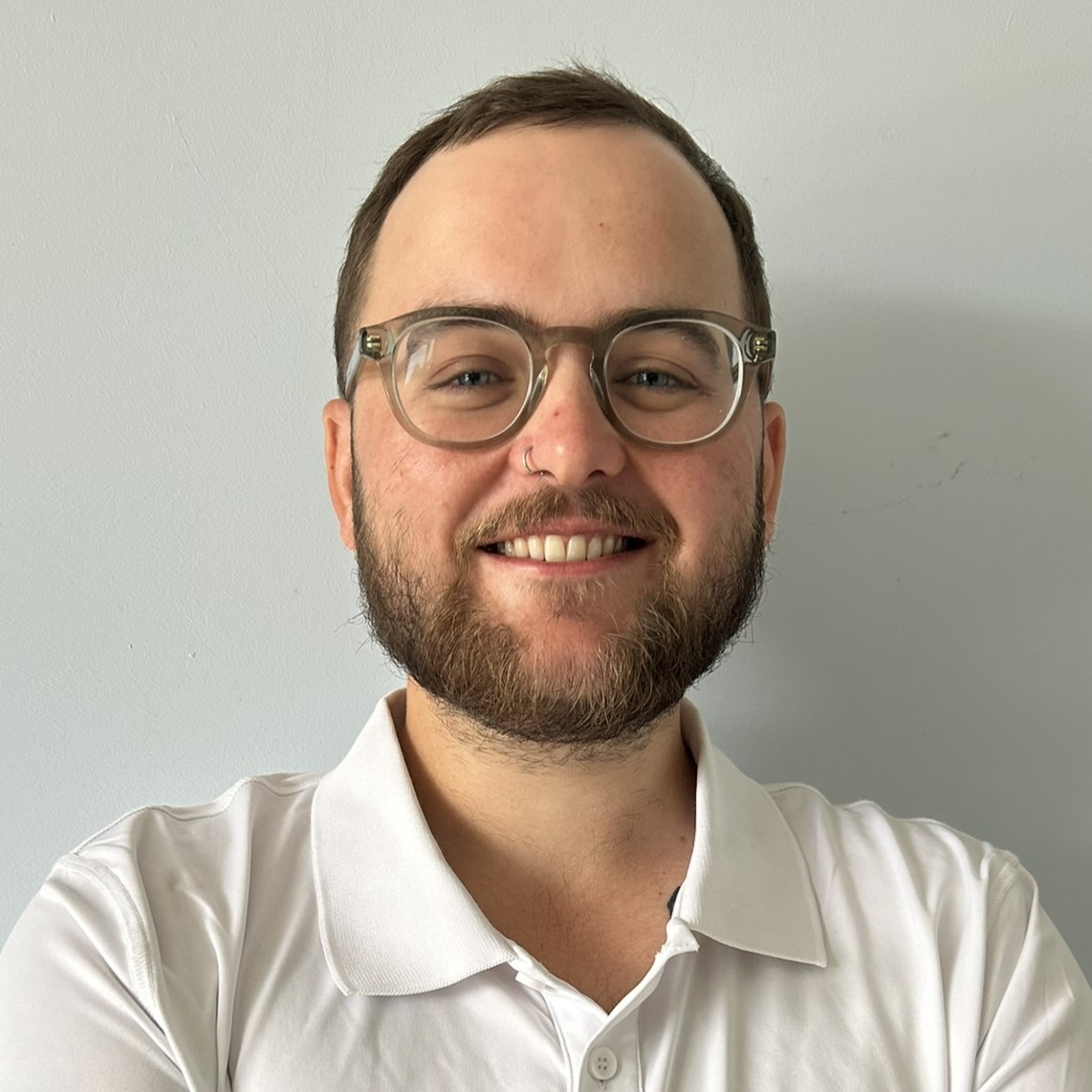 A photo of Ronan - a white man with glasses, brown hair and beard, wearing a white polo shirt and smiling